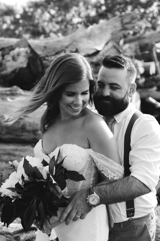 black and white photo of bride and groom cuddling by pile of logs
