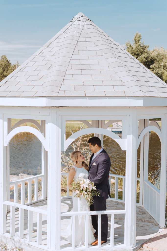 bride and groom standing under white gazebo by the water smiling