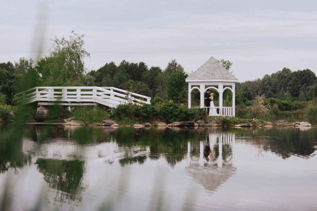 bride and groom standing under little gazebo at Orchard View with reflection in the water