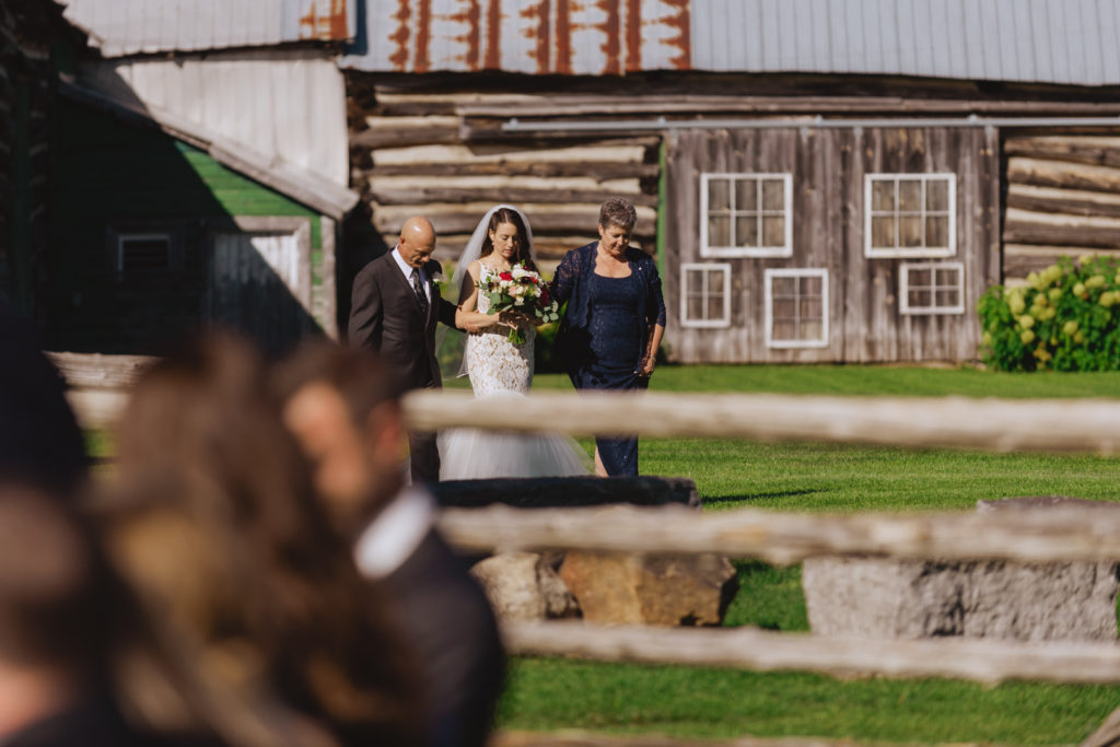 bride walking to the ceremony with her parents with barn behind her