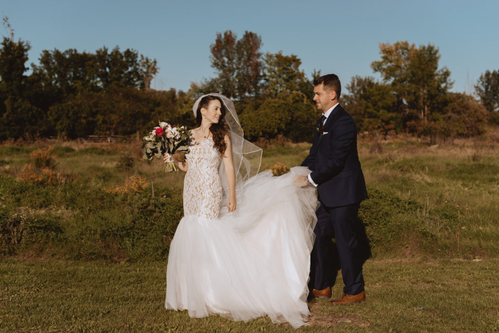 groom helping bride with her train as they walk through a field