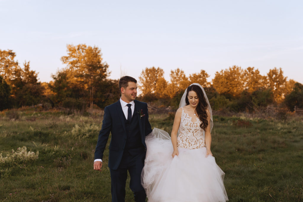 groom holding bride's dress as they walk through a field at sunset