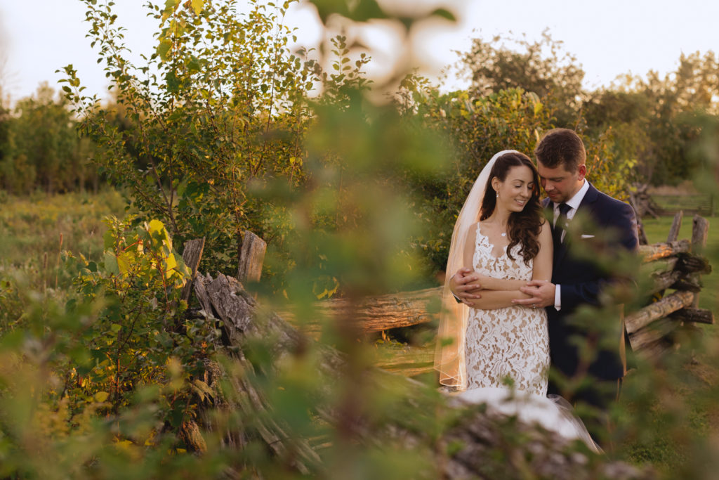 bride and groom cuddling by wooden farm fence at sunset