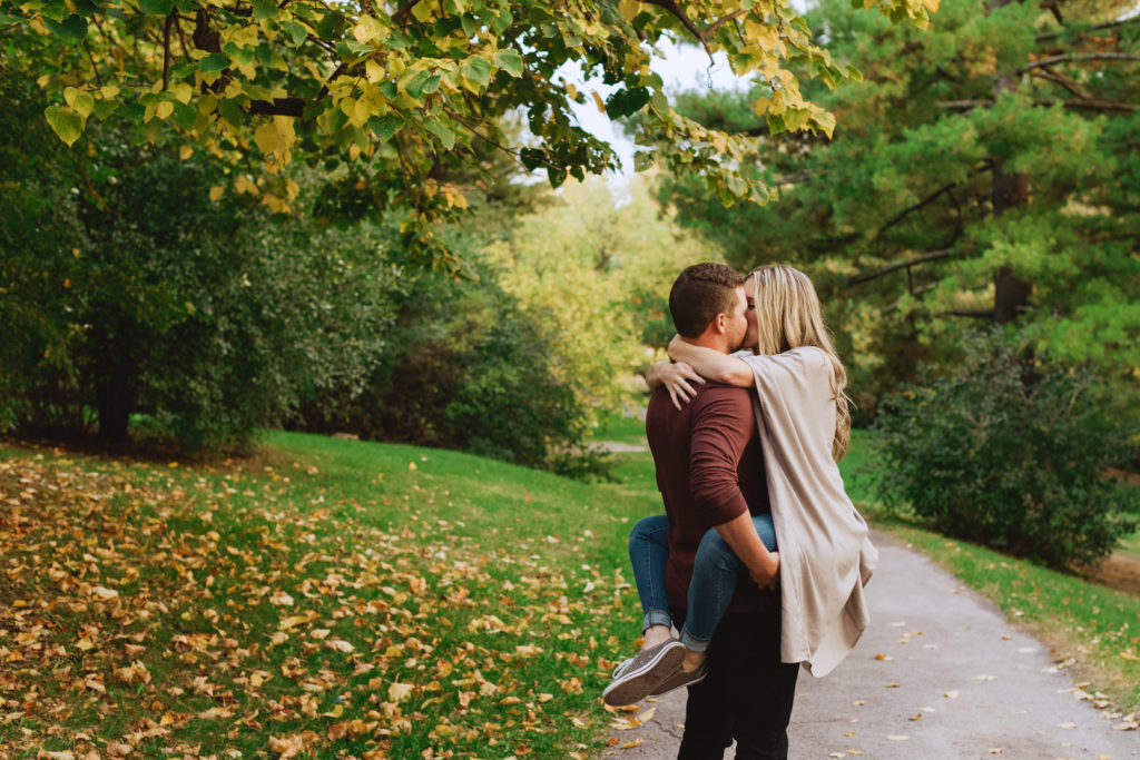 engaged girl with her arms and legs wrapped around her fiancee kissing on gravel path