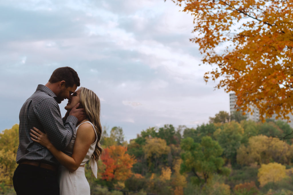 engaged couple kissing with fall coloured trees in the background