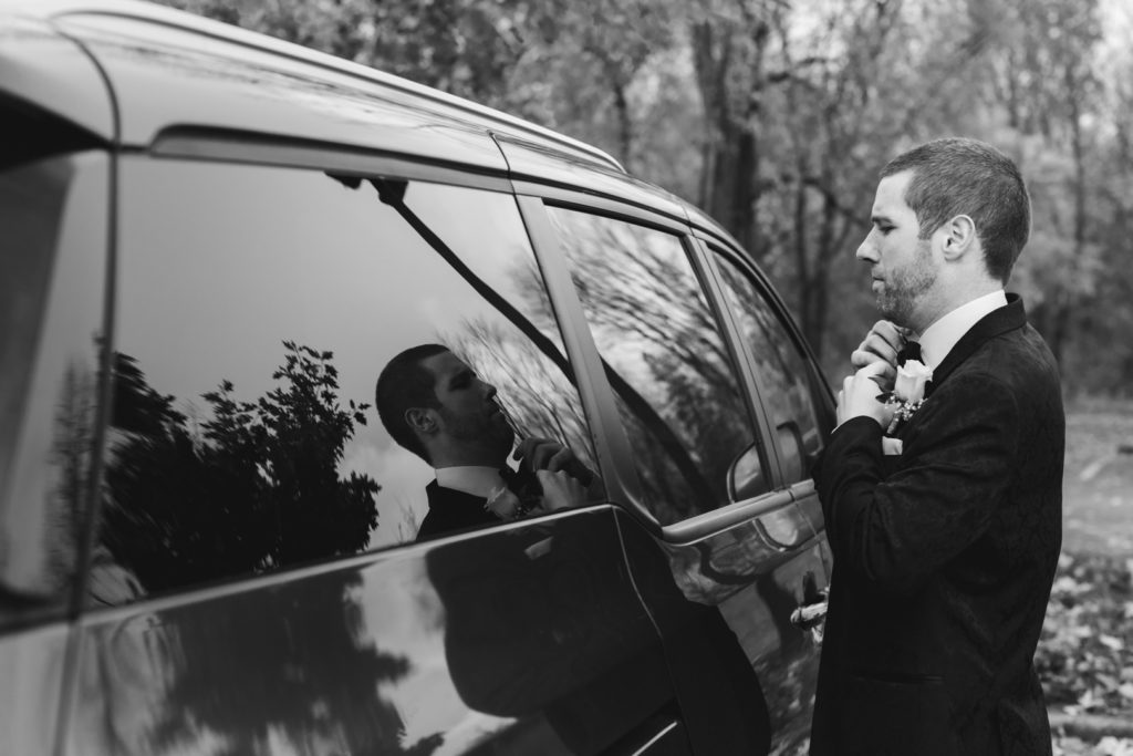 groom adjusting his bowtie in the reflection of a car window