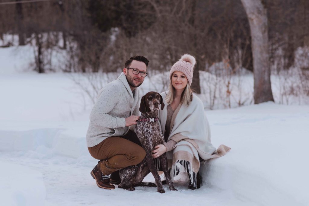 engaged couple smiling with their dog in the snow