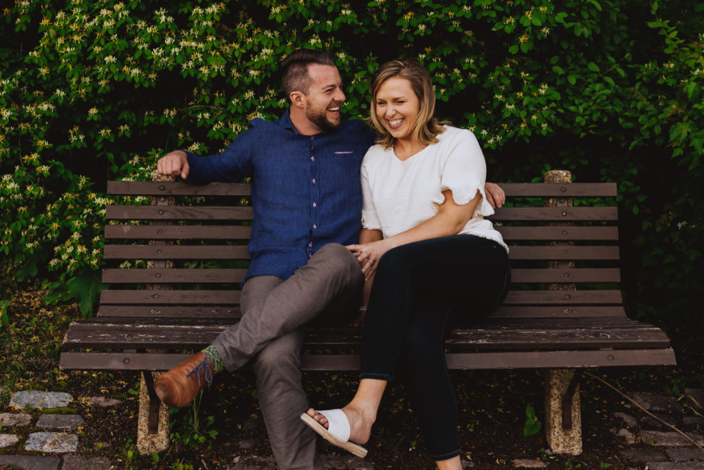 engaged couple sitting on park bench laughing with trees behind them