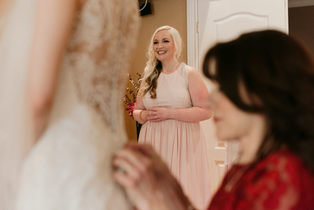 bridesmaid laughing while mom puts on the bride's dress