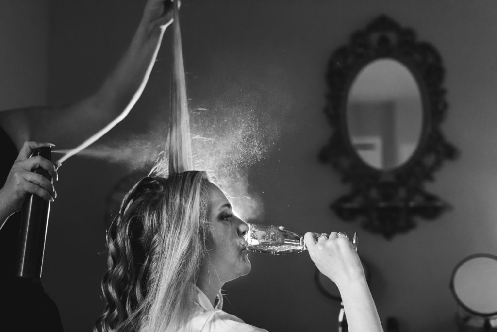 bride drinking champagne while hairstylist sprays hairspray
