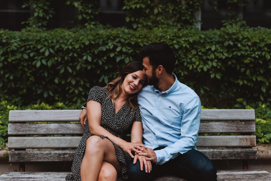 engaged couple sitting on a bench in front of wall with vines