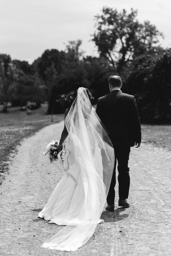 bride and groom walking down dirt trail with cathedral veil blowing in the wind