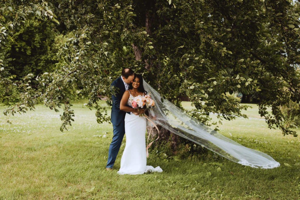 bride and groom underneath tree with wedding veil blowing in the wind