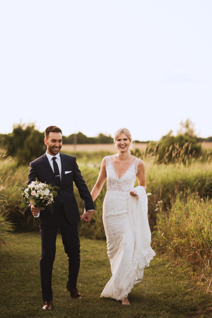 Groom holding bouquet and bride's hand walking through the tall grass at Fields on West Lake