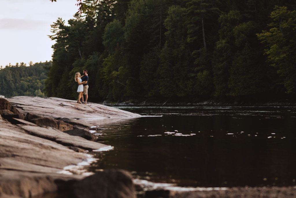 engaged couple standing on rocks by the water at sunset
