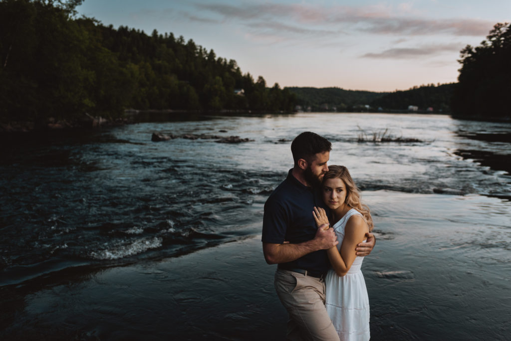 engaged couple cuddling by the water at sunset