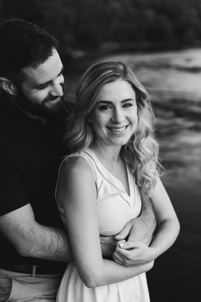 engaged couple cuddling by the water in black and white