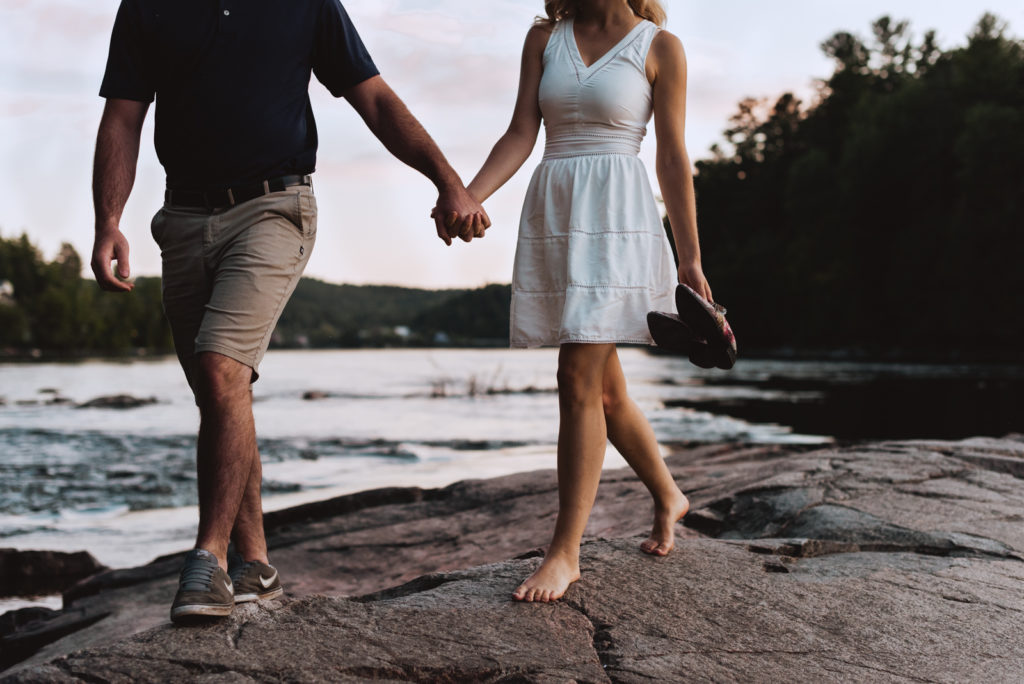 engaged couple holding hands barefoot walking along rocks by the water