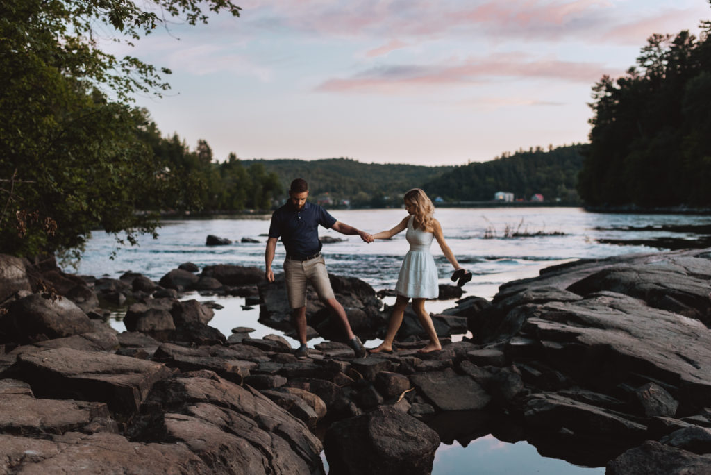 engaged couple walking along rocks by the water at sunset