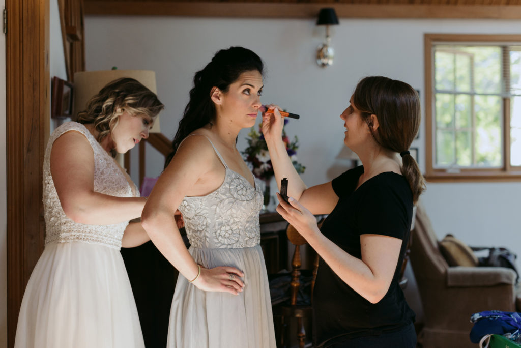 bride helping bridesmaid into her dress while makeup artist touches up her makeup