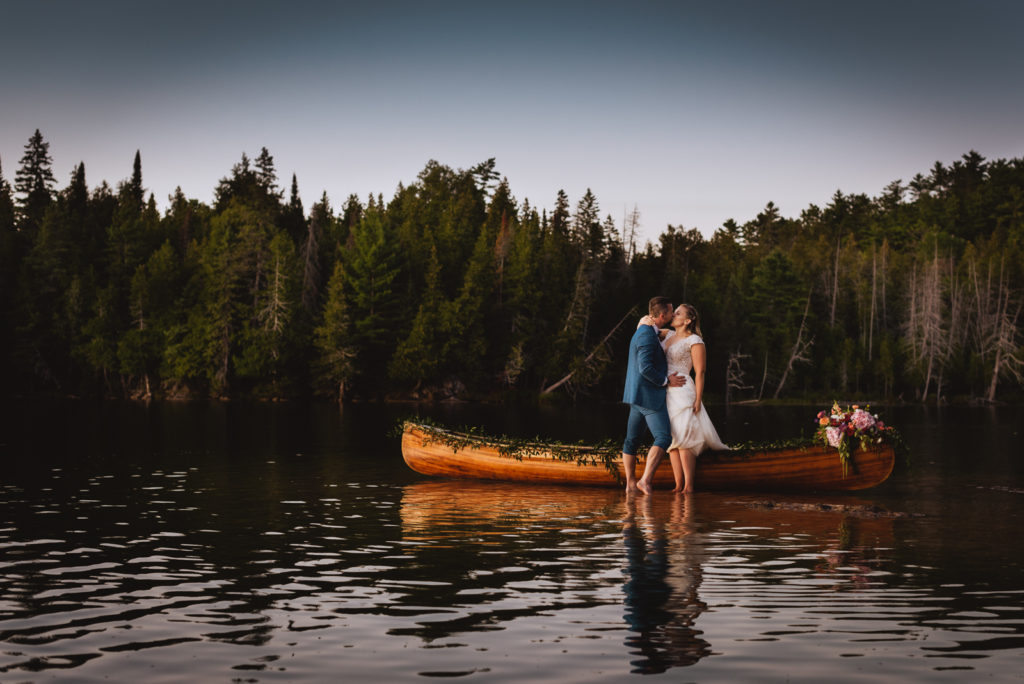 bride and groom standing in the water at sunset by floral canoe
