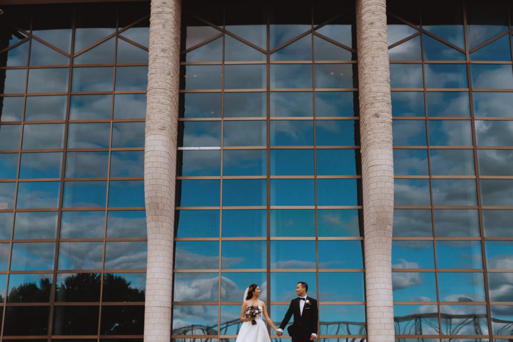 bride and groom holdings hands with large windows reflecting the blue sky