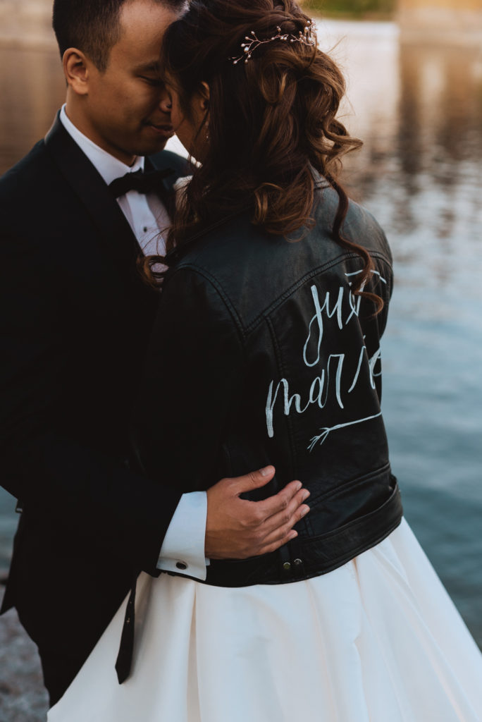 bride wearing the just married leather jacket cuddling with groom by the water at sunset