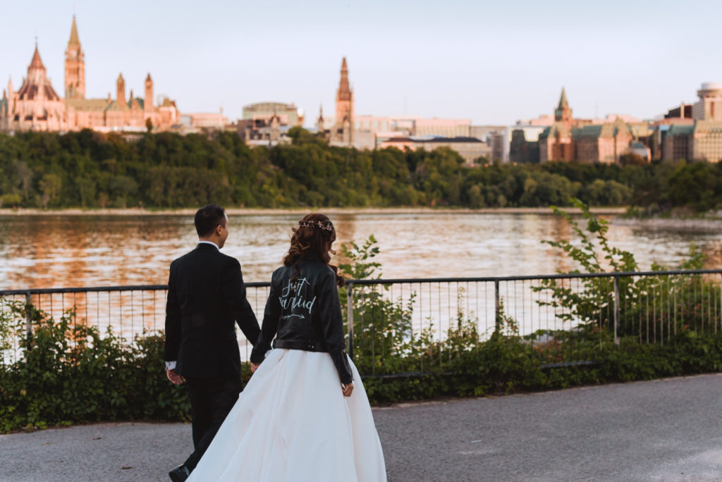 bride wearing just married leather jacket holding groom's hand as they walk along the river overlooking the parliament buildings