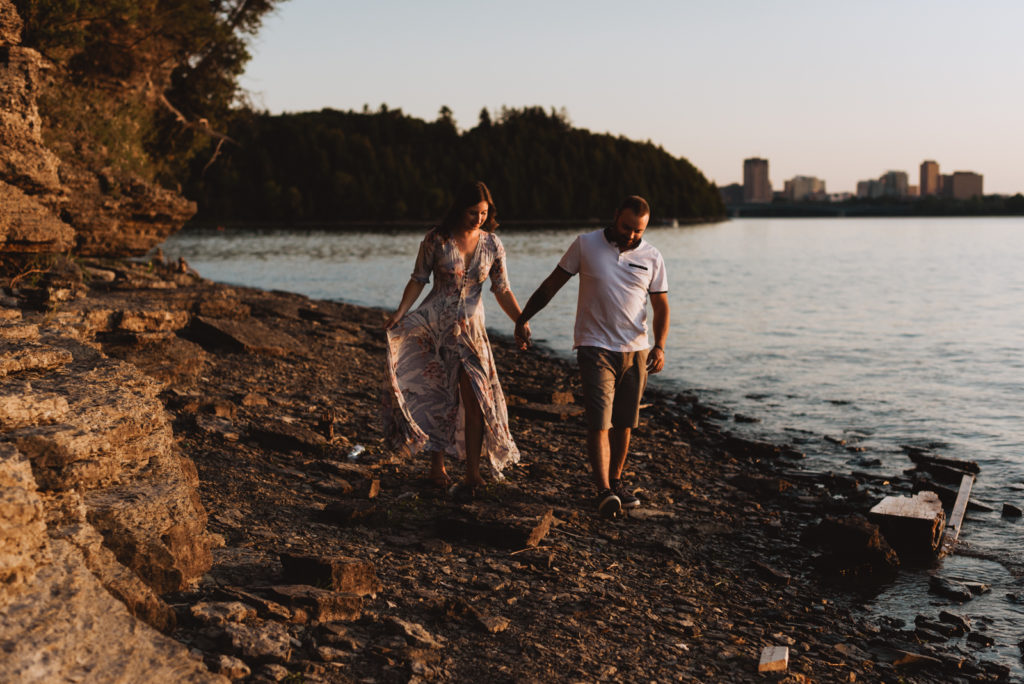engaged couple walking along the river's edge at sunset holding hands