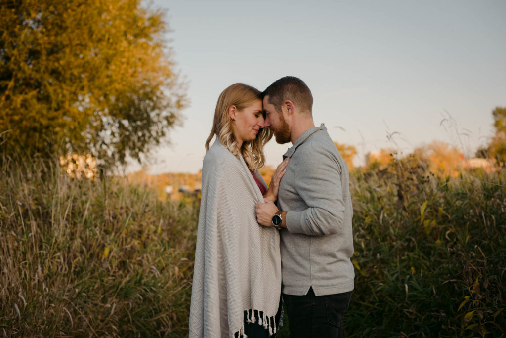 engaged couple with a blanket wrapped around them kissing at sunset in a field