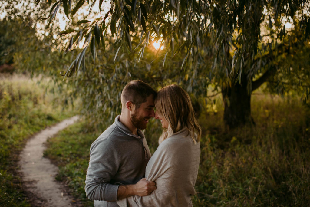 engaged couple with a blanket wrapped around them at sunset under a tree