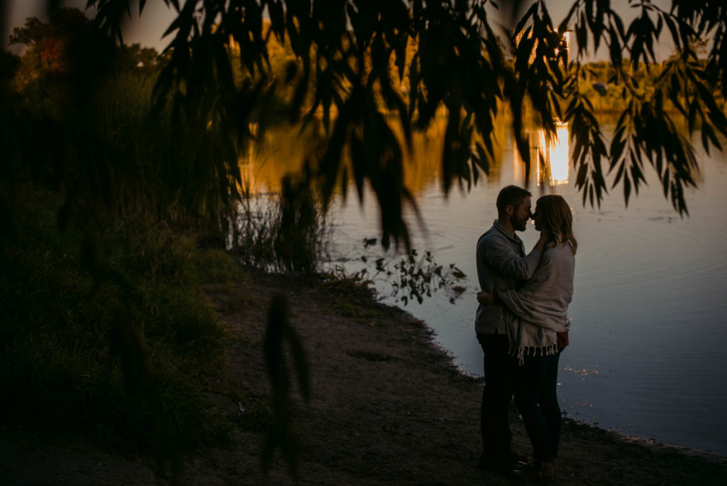 engaged couple cuddling by the water as the sun sets