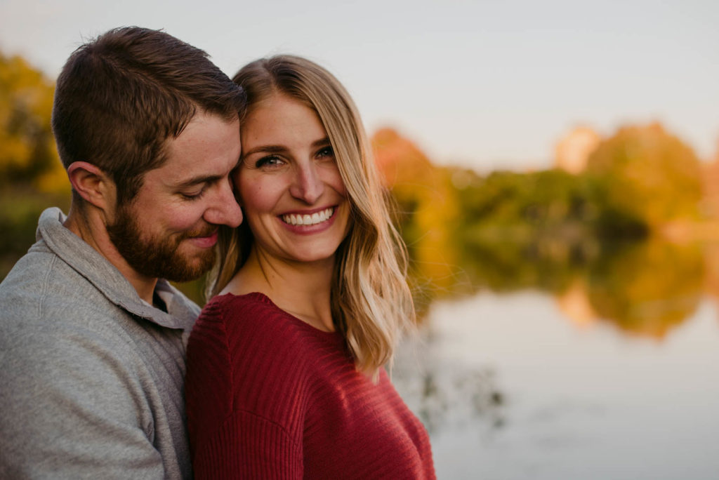 engaged couple smiling and cuddling by the water at sunset