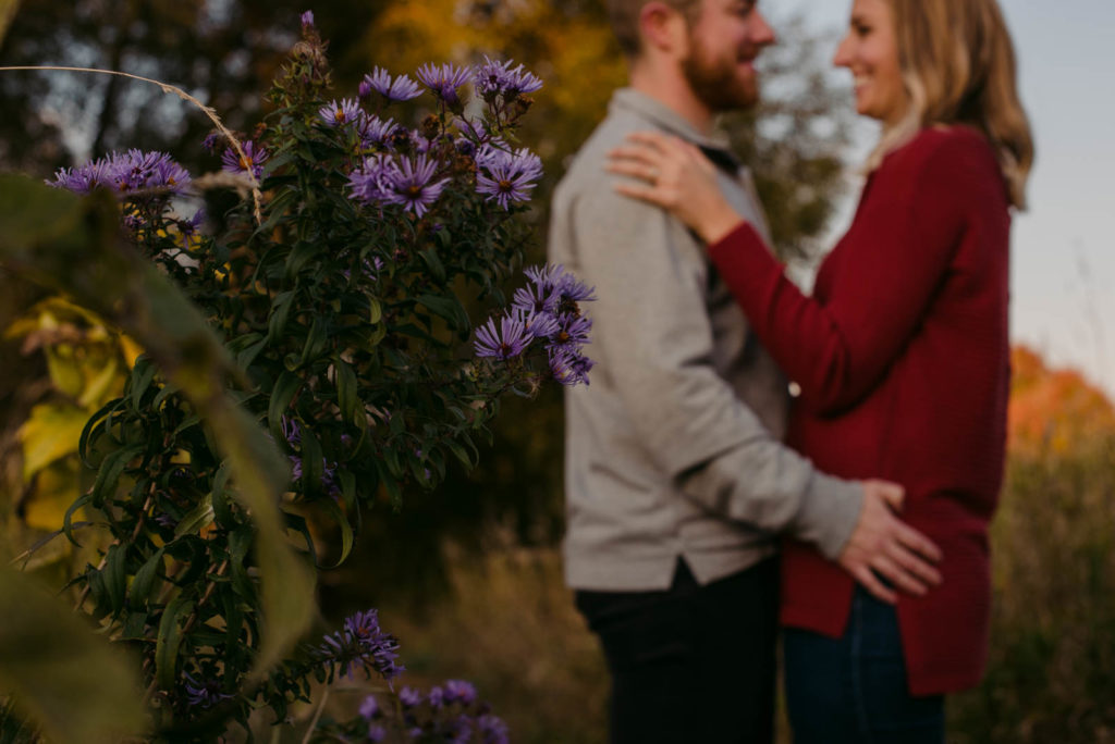 engaged couple cuddling and laughing at sunset with purple flowers