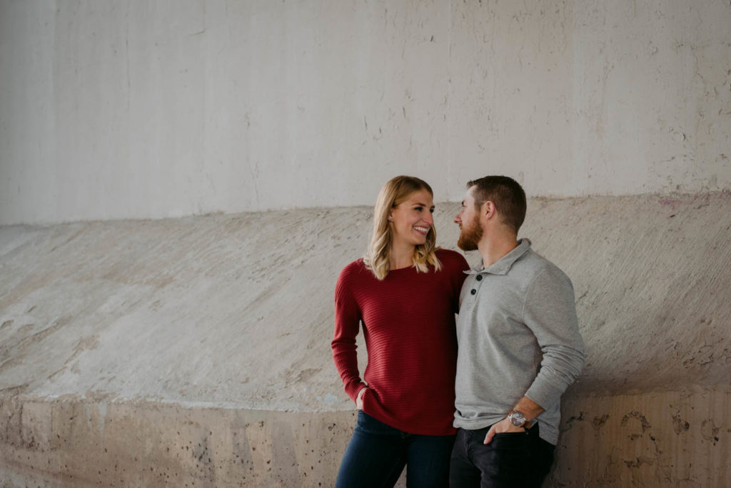 engaged couple smiling with each other underneath a bridge at sunset