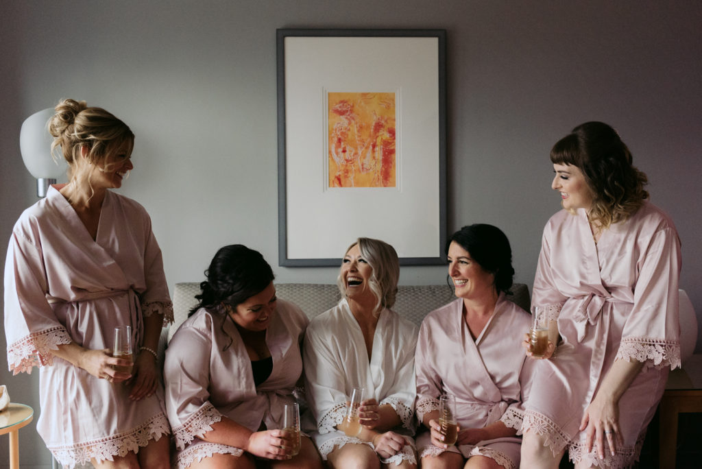 bride and bridesmaids sitting on couch laughing with champagne in robes