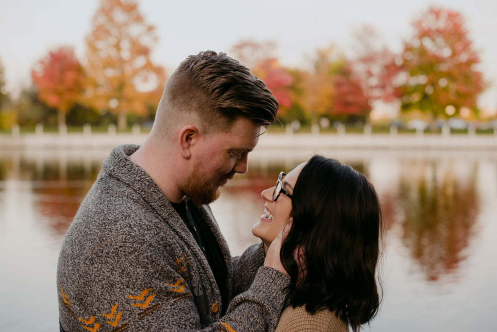 engaged couple cuddling by the water with maple trees reflecting in the water
