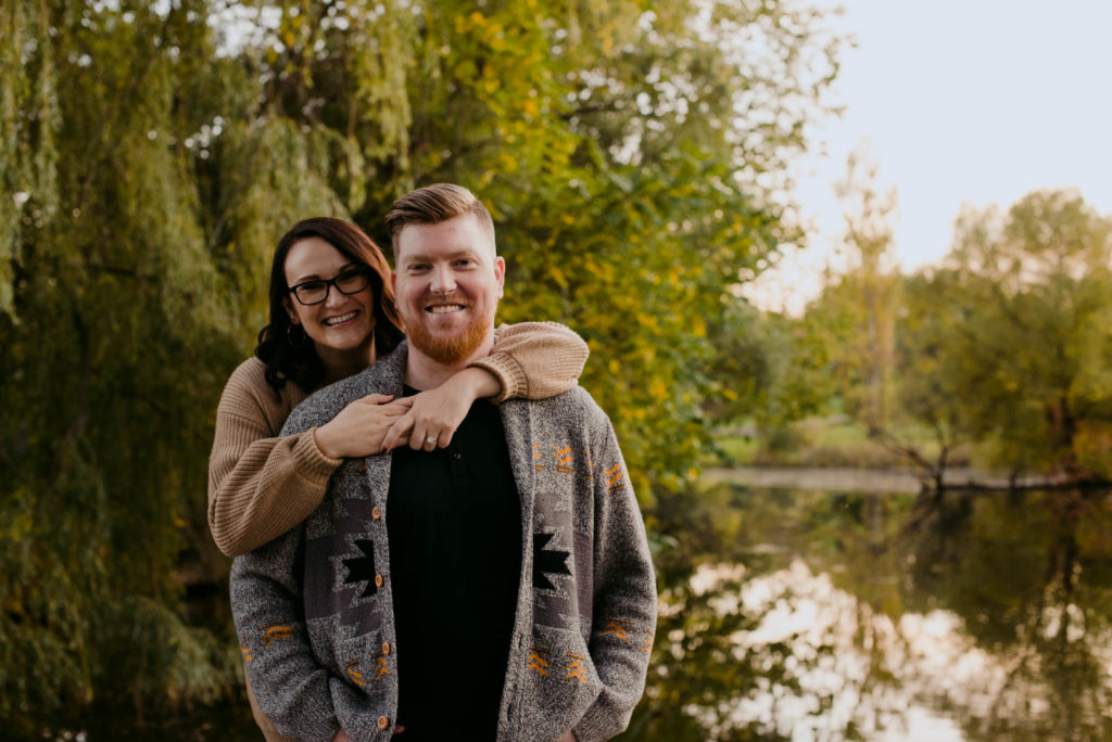 engaged couple in front of willow tree by the water smiling at the camera