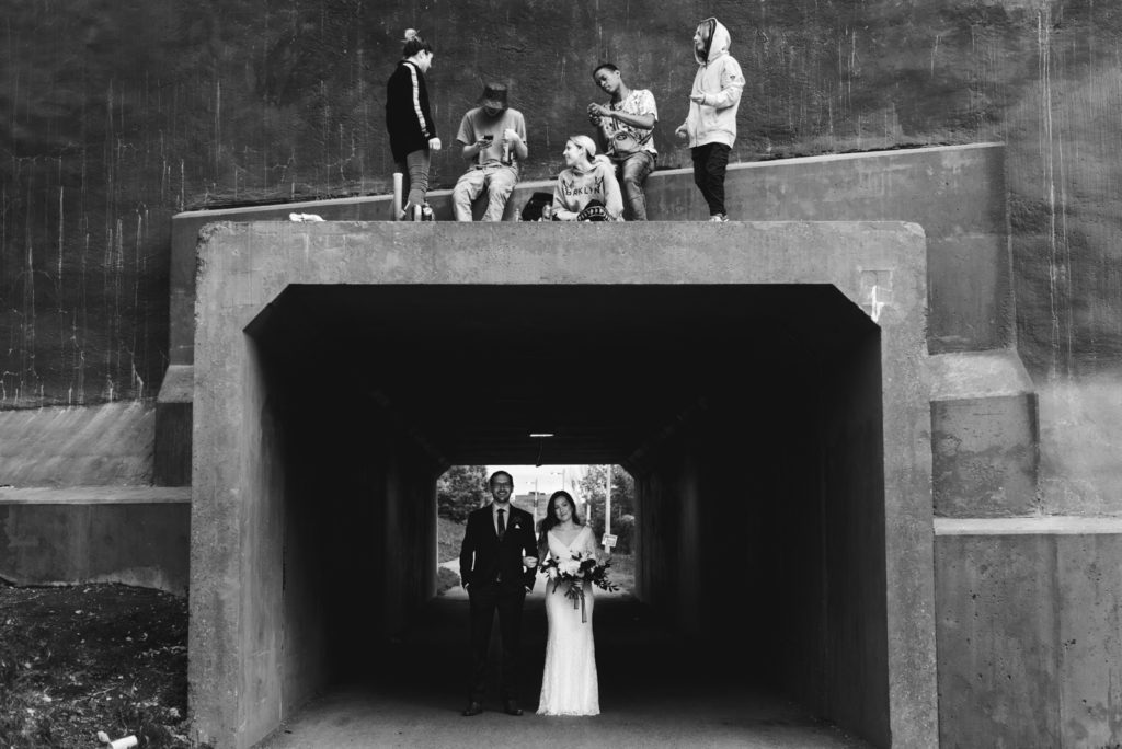bride and groom underneath a bridge with street kids standing above them
