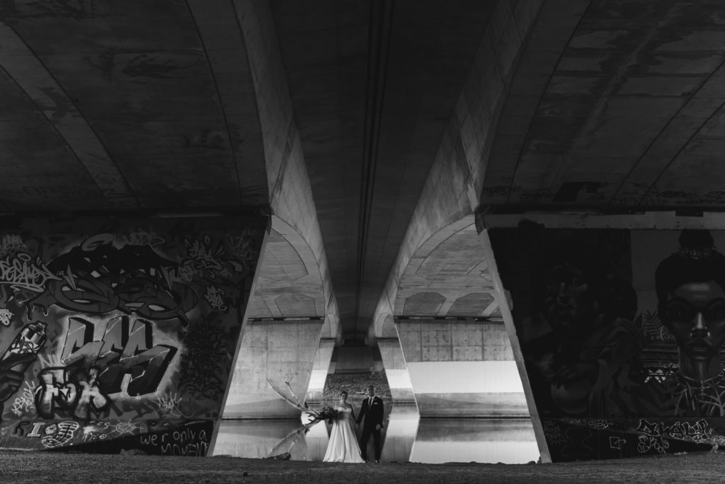 bride and groom standing underneath a bridge with graffiti in black and white