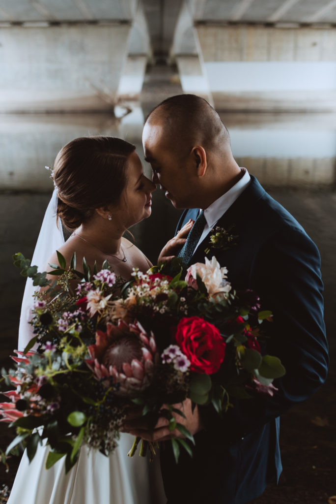 bride and groom cuddling underneath a bridge with wild bouquet