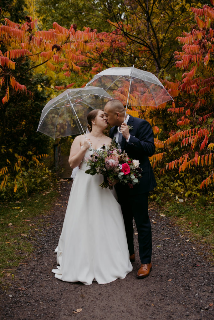 bride and groom standing under clear umbrellas in front of orange sumac trees during fall wedding