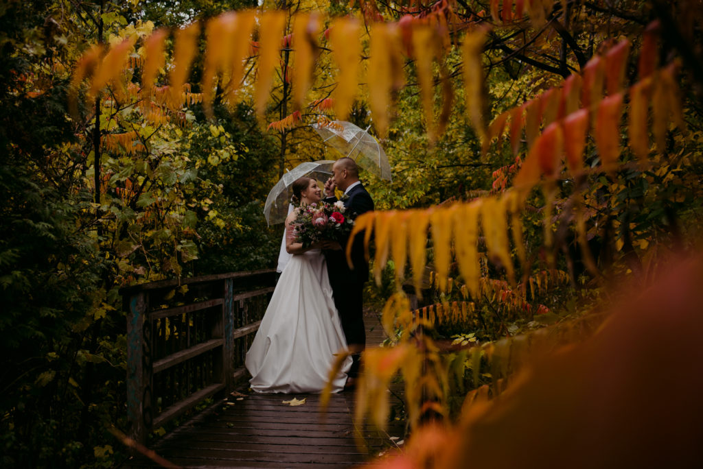 bride and groom standing under clear umbrellas in front of orange sumac trees during fall wedding