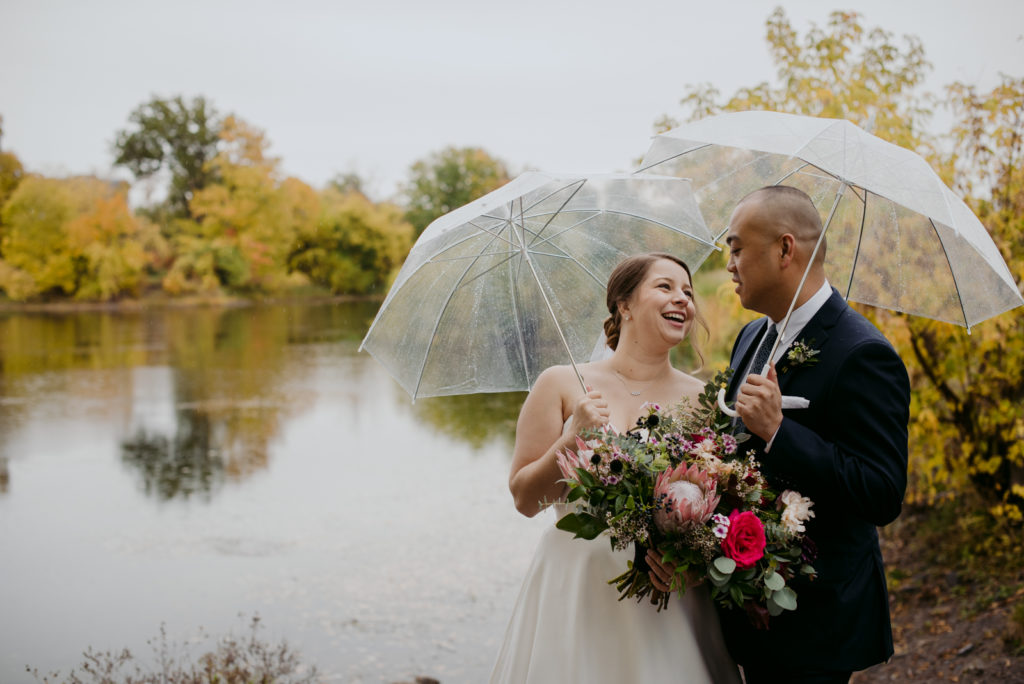 bride and groom laughing underneath clear umbrellas by the water on a rainy day