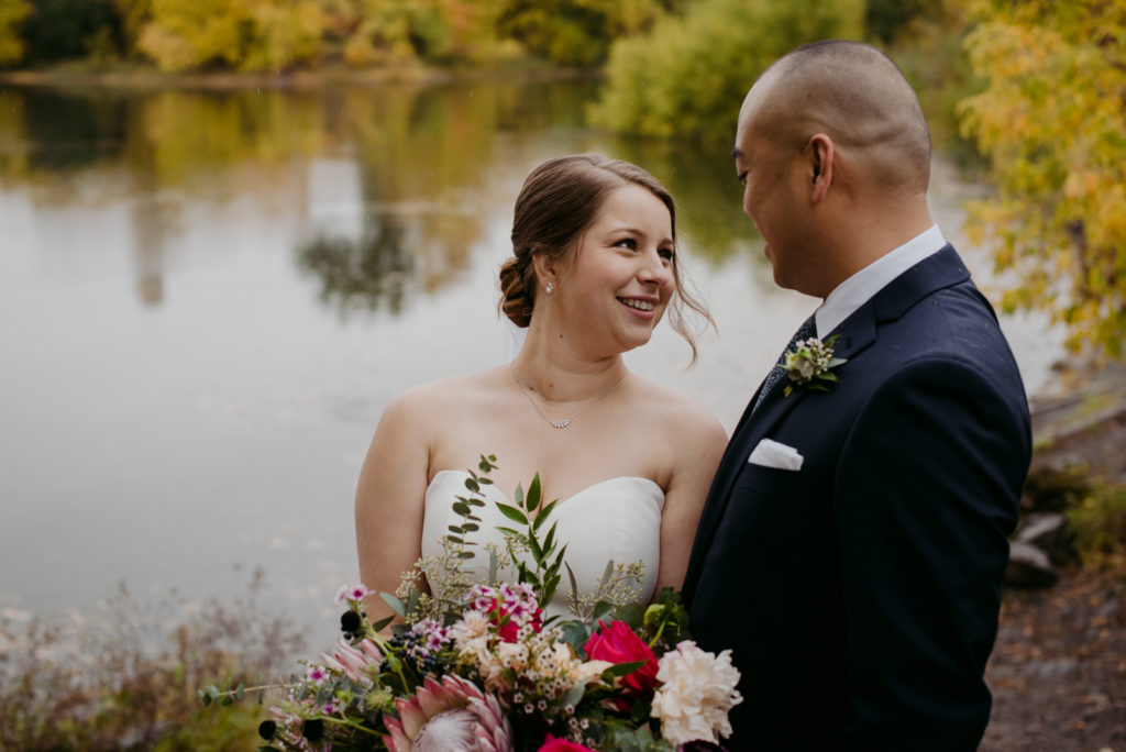bride and groom standing by the water smiling at each other on a rainy fall day