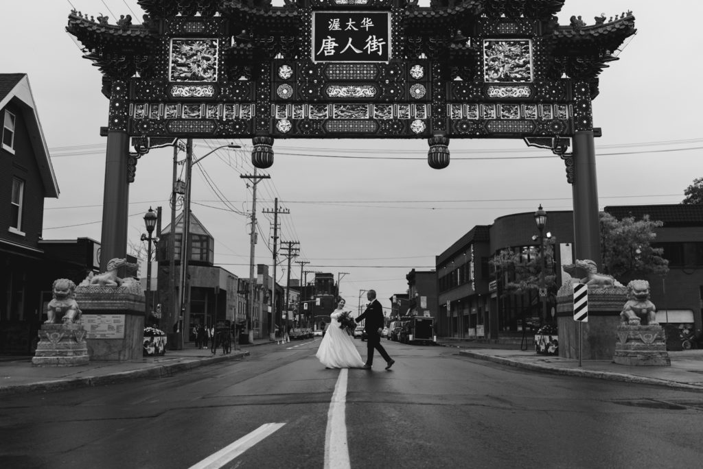 bride and groom walking across the street underneath the Chinatown Royal Gateway in Ottawa