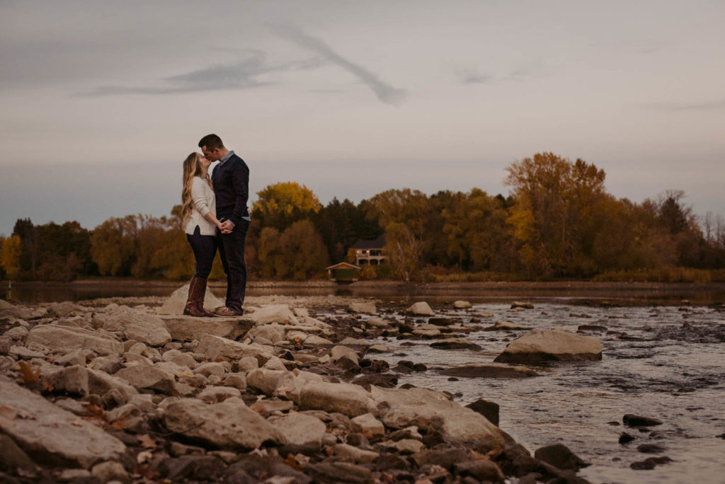 engaged couple kissing on shallow rocks by the water at sunset