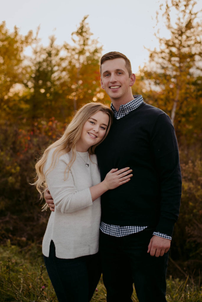 engaged couple cuddling in front of fall trees at sunset