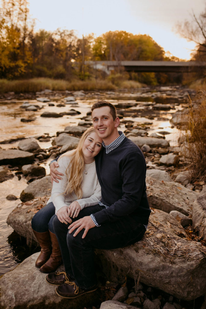 engaged couple sitting on a rock by the water smiling at the camera at sunset