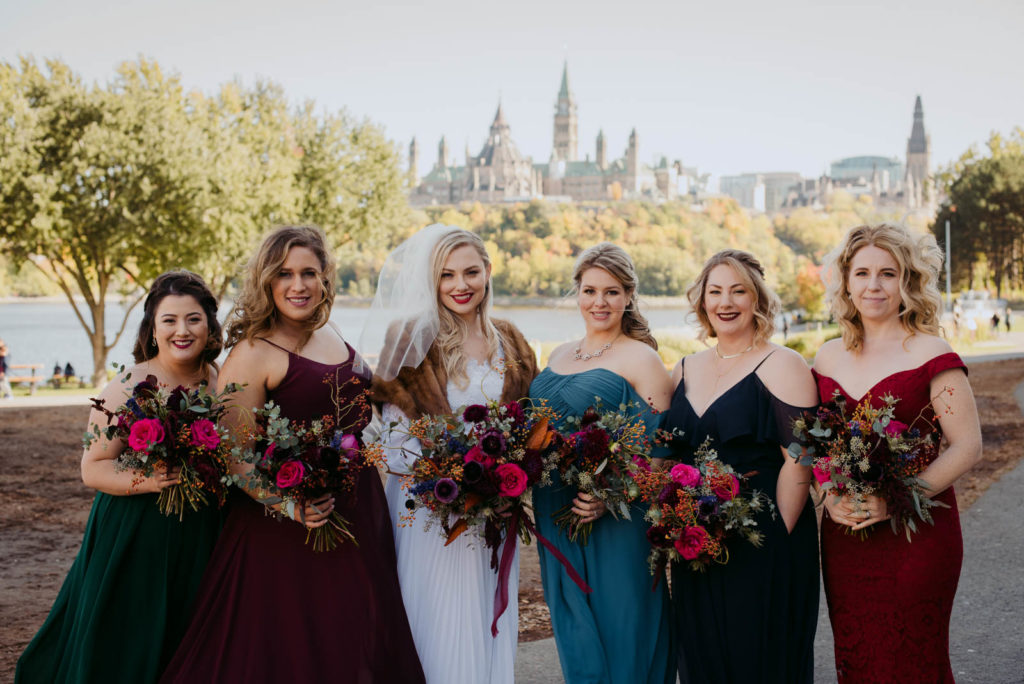 bride wearing fur shawl holding a wild bouquet with her bridesmaids in colourful dresses in front of the parliament buildings